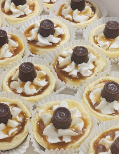 A close-up of mini cheesecakes topped with swirls of whipped cream, drizzled caramel sauce, and chocolate candies placed in the center. The cheesecakes are arranged in white paper liners.