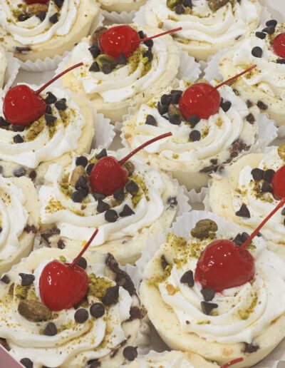 A tray filled with mini cheesecakes, each topped with a swirl of whipped cream, chocolate chips, chopped pistachios, and a bright red maraschino cherry. The desserts are arranged in white cupcake liners.