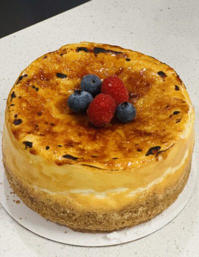 A round, creamy cheesecake with a lightly torched top sits on a white surface. It is decorated with a cluster of fresh berries, including three raspberries and two blueberries at the center. The crust looks crumbly and fully baked.