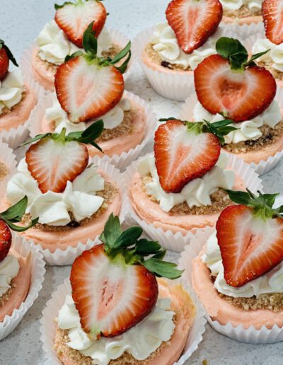 Mini strawberry cheesecakes in white cupcake liners, each topped with whipped cream, a halved fresh strawberry, and a sprinkle of graham cracker crumbs, are arranged on a white surface. The cheesecakes have a pink hue and look freshly made.