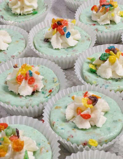 A tray of mint green mini cheesecakes topped with dollops of whipped cream and colorful fruity cereal pieces. Each cheesecake is individually wrapped in white paper cups.