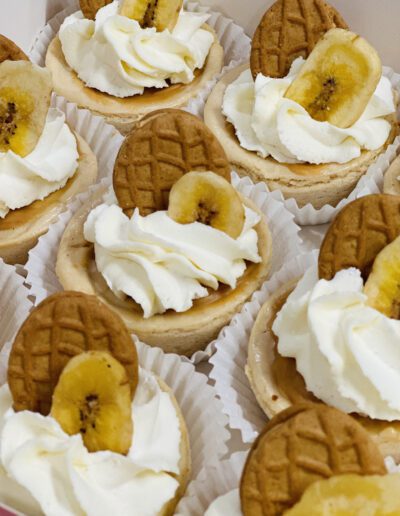 A close-up of several mini cheesecakes, each topped with a swirl of whipped cream, a banana slice, and a waffle cookie, arranged in paper liners.