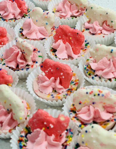 A close-up image of colorful bite-sized treats. The treats feature white and pink frosting with sprinkles and animal-shaped cookies on top, all arranged neatly in white paper cupcake liners. The overall look is festive and playful, with a variety of colors.