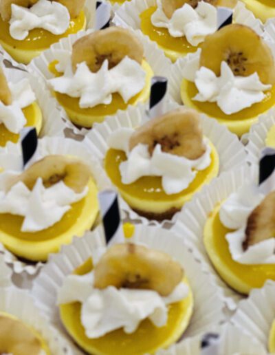 Miniature desserts are arranged neatly in white cupcake liners. Each dessert consists of a yellow base, topped with a dollop of white whipped cream, a slice of banana, and a mini black-and-white striped straw. The overall presentation is vibrant and appetizing.