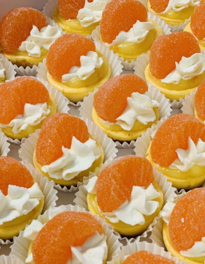 A tray of mini orange-flavored desserts is pictured. Each dessert is topped with a dollop of whipped cream and an orange gummy candy, set on a small, white paper cup. The desserts have a creamy texture and an appealing orange color.