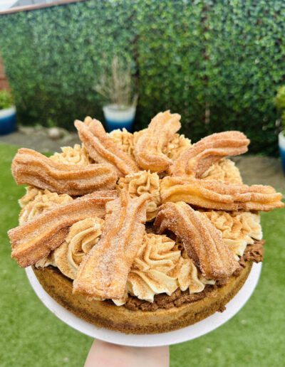 A decadent cake topped with swirls of light brown frosting and artfully arranged churros, dusted with cinnamon sugar. In the background, there is lush green foliage and a manicured lawn, suggesting an outdoor setting.