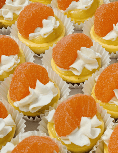 Rows of mini cupcakes topped with a swirl of white frosting and a vibrant, orange gummy candy, all nestled in white paper cups. The cupcakes have a light yellow cream base, creating a visually appealing and colorful dessert display.