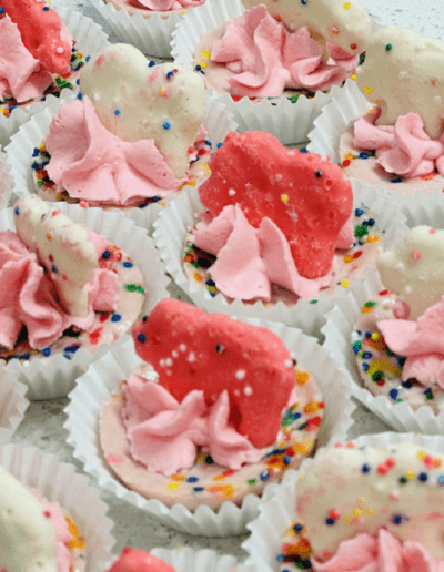 A close-up of colorful mini desserts, each topped with pink whipped cream and animal-shaped biscuits. The biscuits are covered in white or pink frosting with multicolored sprinkles. The desserts are arranged in white paper cups on a white surface.