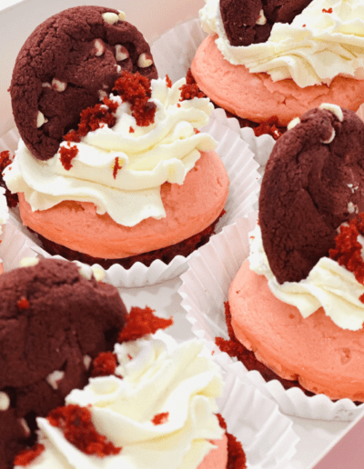 A close-up of red velvet cupcakes topped with swirls of whipped cream, a red velvet cookie, and red crumbs. These cupcakes are arranged neatly in a white box and are in white paper liners. The cupcakes have a pinkish-red cake base and vibrant decorations.