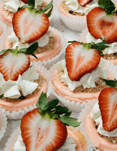 A close-up view of several pink cupcakes topped with whipped cream, crushed graham crackers, and halved fresh strawberries. The cupcakes are arranged in white paper liners, showcasing their decorative and appetizing presentation.